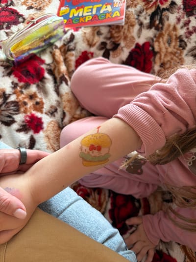 In a close-up shot, a child's arm, adorned with a colorful temporary tattoo of a cartoon character, rests on a patterned blanket. The child is wearing a pink long-sleeved shirt, possibly pajamas, with a subtle bear graphic visible on the fabric. Beside the child's arm lies an open children's book with bold text. A metallic pencil case with a holographic sheen is also present on the blanket. The child's hand, with a black ring on one finger, is held by an adult's hand, suggesting a moment of connection. The overall scene is intimate and appears to be indoors, given the soft lighting and the blanket. The blanket itself features a floral pattern with prominent red and brown elements. The child's skin is fair, and their hair, a light brown, is visible over their shoulder. The image is bright and focuses on the personal interaction and playful elements. There is visible text on the book cover, the words "МЕГА" and "РАСКРАСКА" are discernible, suggesting it's a coloring book. The location context is Valozhyn, Belarus.