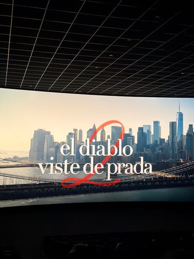 The image displays a large, vertically oriented screen showing a film poster or scene, set within a dimly lit interior space. When properly oriented, the screen presents a sweeping cityscape, identifiable as New York City, with prominent skyscrapers, including one resembling One World Trade Center, under a bright, clear sky, indicating daytime or late afternoon. A body of water and bridge structures are visible in the foreground of the city view.

Overlaid on the urban landscape is bold white text with red outlining, reading "el diablo viste de prada." This Spanish phrase translates to "The Devil Wears Prada," identifying the film. A prominent, stylized red curved element, possibly an 'O' or a design flourish, is integrated with the text.

To the left of the screen, a dark, possibly black, architectural surface features a distinctive grid pattern formed by thin, light-colored lines. This grid extends into the distance, creating a strong sense of perspective and depth within the room or structure. Small, reflective circular points are scattered intermittently along these grid lines. The image captures the viewing experience of this cinematic display, presumably in Paterna, Spain.