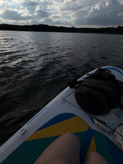 The image shows a view from a paddleboard on a body of water. A dark green backpack and white Crocs shoes rest on the paddleboard. A person's legs are also visible. The water is dark and slightly rippled, and the sky is filled with clouds, next to a forest on the shoreline. The location is presumed to be in the city of Selychka, Russia.