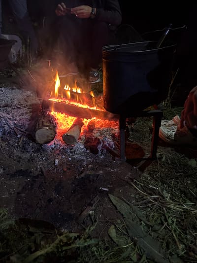 The image depicts a nighttime campfire scene likely in a rural or outdoor setting near Терси, Russia, based on the location request. A black pot hangs over a fire built with logs and kindling, with visible flames and embers. Ash and charred remnants surround the fire. Two people are partially visible in the image. One is wearing a dark jacket, with a hand and watch visible, holding what appears to be a small stick or match. Another person, wearing white shoes, is also visible near the pot and the fire pit. The surrounding environment includes dark vegetation, possibly grass and weeds. The scene appears to be set up for cooking or warmth around the campfire.