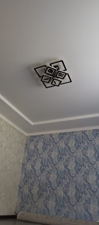 The image shows a modern, geometric ceiling light fixture with black and white square-shaped elements, suspended from a plain white ceiling. The ceiling has decorative white molding along the edges where it meets the walls. The walls are covered in wallpaper. The wall on the left has a light-colored, textured wallpaper with a marbled or abstract pattern. The wall on the right has a darker wallpaper with a blue and gray marbled or abstract pattern. A rolled-up carpet with a patterned edge is visible on the floor in the lower right corner of the image. The scene appears to be an interior of a room, likely a living room or dining area, in Barskoon, Kyrgyzstan. No people are present in the image.