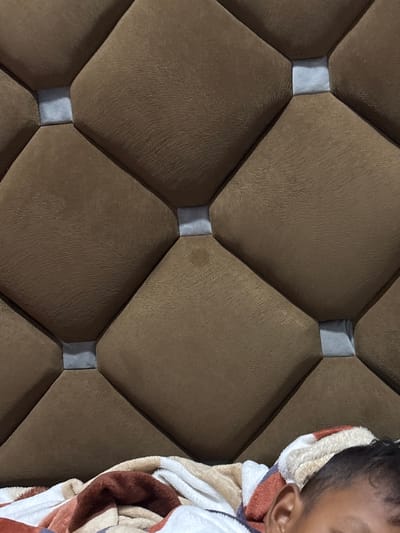 The image shows a close-up of a brown upholstered headboard with a diamond-stitched pattern. Interspersed between the diamond shapes are small, light gray fabric squares. In the lower right portion of the image, a person's ear and the back of their head are visible, resting on a multi-colored blanket. The person appears to be sleeping or resting. The scene suggests a bedroom setting, likely in a home. The location is identified as Geulanggang Teungoh, Indonesia.