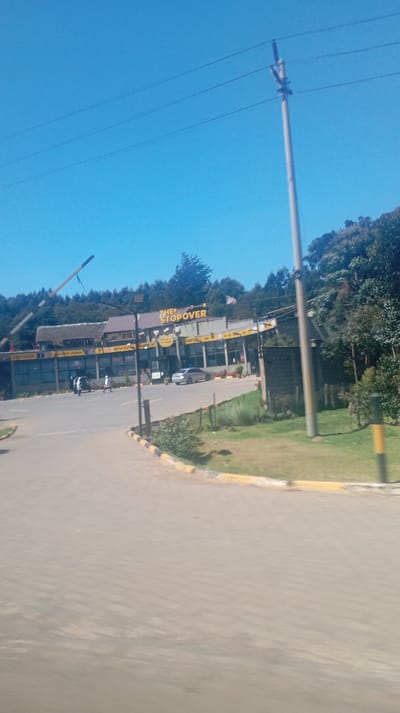 The photo depicts "The Stopover" establishment with a building housing various services like a restaurant and lounge. A road leads to the entrance where a couple of people are seen walking. The scene is bright under a clear blue sky, with greenery surrounding the building, situated on a hill. A power pole stands prominently on the right side of the image, adding to the suburban environment.