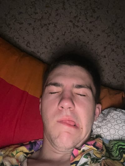 The image shows a close-up, overhead view of a young adult male lying down, seemingly asleep or drowsy. He has his eyes closed and his mouth is slightly open with his tongue protruding, giving him a humorous or disheveled appearance. He is wearing a colorful, patterned shirt. The man is lying on bedding that includes a red and orange surface and a white patterned fabric. The background appears to be a dark, textured surface, possibly a ceiling or wall with a subtle, speckled pattern. There is no information about the location in the city of Ponebel, Ukraine.