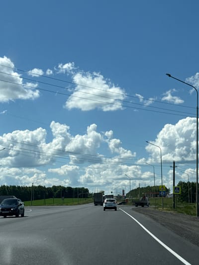 This is a vertically oriented, eye-level, medium shot of a highway in Kendektamak, Russia, under a bright, cloudy sky. The sun is high, suggesting it is midday. The road is paved with an asphalt surface and has a white line marking the right edge. To the left of the road, there is a grassy verge with trees and poles with power lines. There are several cars and trucks on the road, indicating traffic flow. A pedestrian crossing sign is visible on the left side of the road. The sky is a vibrant blue, with large, fluffy white clouds scattered across it. The overall scene conveys a sense of a typical day on a highway in a rural or suburban area. There are no visible emotions.