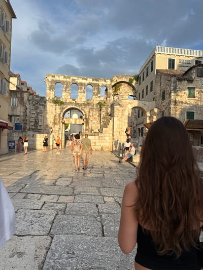 The image depicts a sunny day in Split, Croatia, with the setting sun casting long shadows across an ancient stone courtyard. The architecture suggests a historic location, possibly within the walls of Diocletian's Palace. In the foreground, a person with long brown hair is seen from behind, looking out at the scene. Several other individuals are scattered throughout the courtyard, some walking, others resting or standing. One man in a light green t-shirt and shorts stands out, along with a couple walking together. The stone pavement is worn and irregular, indicative of its age. A partially visible building with the word "HOTEL" and "RESTAURANT" on its facade can be seen in the upper left corner. The sky is partly cloudy, with a mix of blue sky and white clouds. The warm light suggests it is late afternoon or early evening.