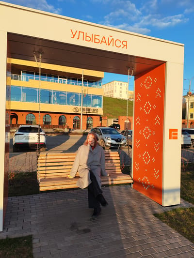 The photograph captures a woman sitting on a wooden swing that is part of an outdoor structure located in the city of Средний Постол, Russia. The structure features a white frame with orange accents. The word "УЛЫБАЙСЯ" (which translates to "smile") is displayed in orange text along the top of the frame. One side of the structure has a decorative orange panel with geometric patterns.
The woman has reddish-blonde hair and is wearing a gray coat over dark clothing. She sits with her legs crossed, looking slightly to the left of the frame.
In the background, a multi-story building is visible, along with several parked cars. The sky above is a gradient of blue and light orange, indicating either sunrise or sunset. The ground is paved with gray bricks, and there are patches of grass nearby. The overall setting appears to be a public space or park-like area.