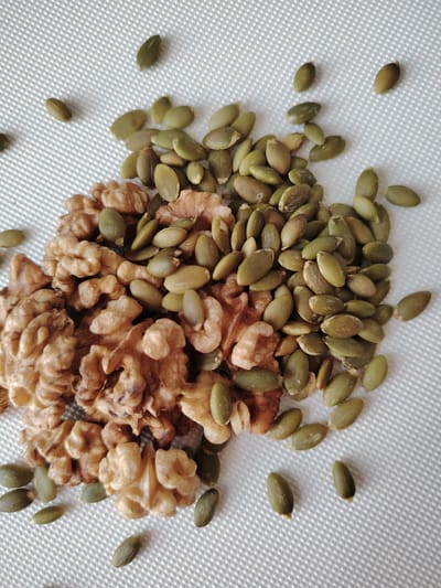 A pile of mixed walnuts and pumpkin seeds is scattered on a white, textured surface. The walnuts are broken into irregular pieces, showing their light brown, wrinkled texture. The pumpkin seeds are an olive-green color, mostly intact, and oval-shaped. Some individual seeds are scattered around the main pile, with a few larger clusters of seeds and walnuts also visible. The lighting suggests an indoor setting, possibly a kitchen or a studio, with no indication of time of day or weather. There is no visible text in the image. The scene is a simple, still life composition focused on the food items.