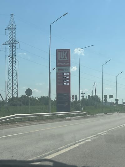 The image depicts a roadside scene in Risovyi, Russia, on a clear day, likely in the late morning or early afternoon. The foreground is dominated by a gas station sign with the logo "ЛУКОЙЛ" prominently displayed. Below the logo, a series of numbers are visible, likely representing fuel prices, though the specific values are obscured.

To the left of the gas station sign, a large, lattice-style electrical tower stretches towards the sky, crisscrossed with power lines. Several streetlights are also visible throughout the scene, some with curved arms extending over the road. In the background, trees line the side of a paved road, which appears to be a highway. The road has a yellow dividing line and a white edge line, and a concrete barrier runs along the side.

Further in the background, several traffic signs and smaller utility poles can be seen. The sky is a bright, clear blue with a few wispy clouds, indicating fair weather. The overall impression is of a typical, functional roadside environment in a rural or semi-rural setting. There are no people visible in the image.