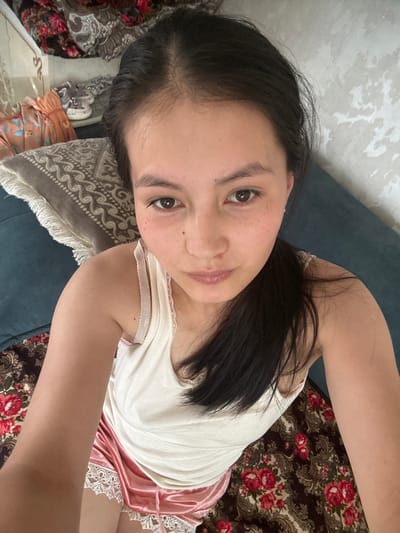 A young woman with dark hair pulled back into a ponytail is seen from a high-angle perspective, looking directly at the camera. She is wearing a white tank top and pink satin shorts with lace trim. She is sitting on a couch with a patterned throw blanket behind her. The environment appears to be indoors, likely in a residential setting, possibly in the city of Жыгач-Коргон, Kyrgyzstan, based on the provided context. The background shows a textured wall and various fabric items, including a patterned cushion. The woman has light skin with freckles across her nose and cheeks.