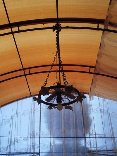 A close-up, low-angle shot shows a decorative outdoor chandelier hanging from a vaulted ceiling structure. The ceiling is made of an orange, translucent fabric stretched over a black metal frame. A section of white, lace-trimmed curtain is visible on the right, partially obscuring the orange fabric. The chandelier is black, featuring a circular frame with decorative elements resembling leaves or flowers. Three light bulbs are visible, hanging downwards from the center of the chandelier. The background is a pale blue, likely depicting the sky, viewed through a translucent, vertically ribbed material, possibly a window or screen, with a patterned lace overlay. Horizontal black bars are visible across the background. The scene appears to be an outdoor or semi-outdoor covered area, such as a patio or gazebo, in Krupiitski sel'ski Savet, Belarus.