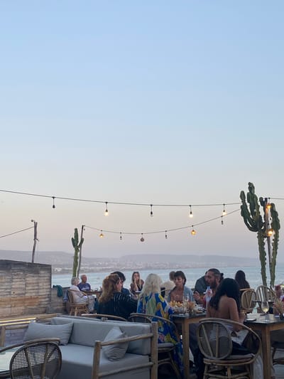 An outdoor dining area overlooks a coastal city in Morocco, likely Taghazout, at dusk. Several tables are occupied by groups of people eating and conversing. The scene is illuminated by string lights and the soft light of the setting sun. Tall, spiky cacti stand as decorative elements. The architecture features wooden walls and rattan furniture. The sea is visible in the background with a hazy coastline.