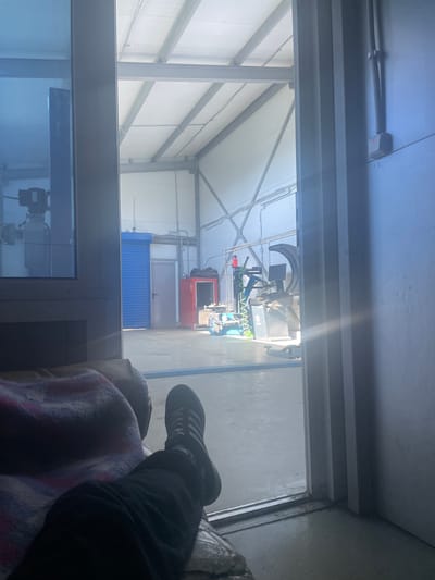 The image depicts the interior of a workshop or garage, likely a tire shop, as suggested by the tire mounting machine visible in the background. The scene is viewed from inside the workshop, looking out through an open doorway or large window. In the foreground, a person is seen relaxing on a couch or chair, covered with a blanket. Their legs are visible, wearing black trousers and black athletic shoes with white stripes. The workshop itself appears spacious with a high ceiling supported by metal beams. Various pieces of equipment, including what seems to be a car lift and a red cabinet, are present. The floor is concrete and appears wet in places. The lighting suggests it is daytime, with sunlight streaming in through the opening and reflecting off surfaces. The location is identified as the city of Kirishi, Russia.
