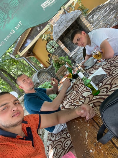 In Sushina, Bulgaria, three young men are seated at an outdoor table, likely at a restaurant or cafe. The man closest to the camera, wearing an orange polo shirt, is taking a selfie. To his right, another man in a teal shirt is holding a piece of food and looking at the camera. The third man, in a white t-shirt, is seated further back, holding a glass of beer and looking towards the camera.

The table is covered with a patterned tablecloth and displays several items, including two glasses of beer, an unopened green bottle of beer, and a bottle of what appears to be wine. There are also plates, a napkin, and a black bag on the table. The background reveals a rustic setting with stone walls, wooden structures, and lush green trees, suggesting an outdoor seating area surrounded by nature. The overall ambiance appears relaxed and casual, as if the men are enjoying a meal or drinks together.