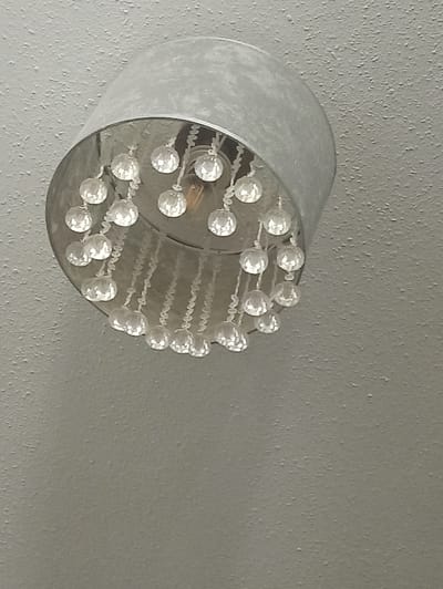 A close-up, upward-angle shot shows a light fixture with a cylindrical, brushed metal shade, attached to a textured, light gray ceiling. Dangling from the inside rim of the shade are multiple strands of clear, faceted crystal beads. The beads are arranged in concentric circles and rows, with individual beads strung together. A faint glow emanates from a single light bulb visible within the fixture. The background consists entirely of the textured ceiling. The image appears to be taken indoors, possibly in a residential or commercial space. There is no information to determine the specific location within Freiburg im Breisgau, Germany.