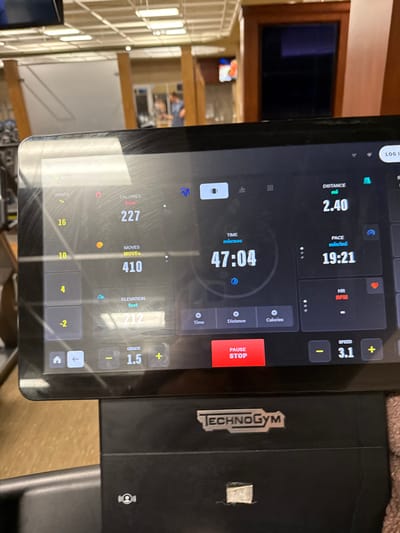 The image displays a close-up of a Technogym treadmill's digital console, which is smudged with visible streaks and fingerprints. The screen shows active workout data: Time elapsed is 47 minutes, 4 seconds; Distance covered is 2.40 miles; Pace is 19 minutes, 21 seconds per mile; Calories burned are 227 kcal; "Moves" count is 410; Elevation gained is 212 feet; Current Grade is 1.5%; and Current Speed is 3.1 mph. The Heart Rate (HR) field displays a dash, indicating no active reading. "WEDNESDAY" and "Apple Watch" integration are partially visible at the top left. Below the screen, the "TECHNOGYM" logo is present on the treadmill's black plastic casing. The background, largely out of focus, reveals an indoor fitness center environment with overhead fluorescent lighting reflected on the screen. Wooden architectural elements, possibly support pillars or door frames, are visible. A blurred individual wearing a light-colored shirt is discernible in the middle-left background, seemingly using other gym equipment. A television screen is faintly visible in the far upper-right background. The scene is located within a fitness facility in Plainsboro Township, United States.