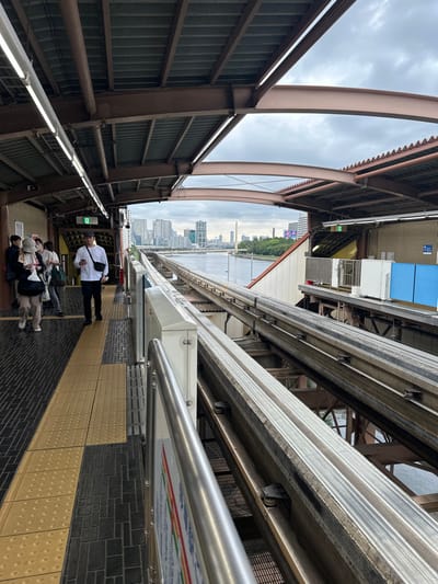 The image displays an elevated monorail station platform, specifically identified as Tennozu Isle Station in Shinagawa Ward, Tokyo, Japan. The perspective looks diagonally across two parallel monorail tracks towards a canal and the distant urban skyline under an overcast sky.

The station platform features black and dark grey patterned floor tiles with a prominent yellow tactile paving strip running along its length. Above, a brown steel support structure holds a corrugated metal roof, illuminated by multiple fluorescent strip lights.

Approximately five to seven individuals are visible on the platform. In the mid-ground, a male in a white long-sleeved shirt, dark pants, and a dark sling bag walks towards the left, focused on a mobile phone in his hands. To his left, a female wearing a light-colored hat and dark jacket, carrying a bag, walks in the same direction, looking downwards. Other individuals are partially obscured in the background, appearing to be waiting or moving along the platform.

Two sets of elevated monorail tracks, constructed as robust concrete or metal guideways with steel running rails, extend into the distance. A metal railing and a modular white barrier system line the platform's edge adjacent to the tracks. Beneath the tracks, a calm, grey body of water, a canal, is visible. Across the canal, a dense city skyline of high-rise buildings and a distinct bridge is apparent against the cloudy sky. A narrow band of green trees lines the far bank of the canal. Additional station elements include a green "Exit" sign on a platform wall and various white and blue utility panels on the opposite side of the tracks.