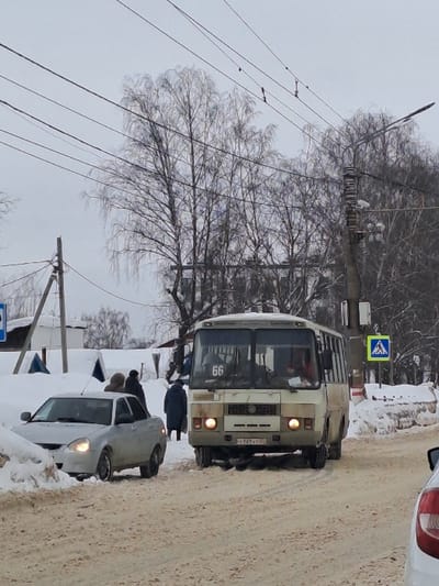 A street scene in winter features a tan bus with "66" displayed on its front, positioned perpendicular to the camera's view. A white car sits close by, partially obscured by snow. Overhead, a web of power lines intersects the sky, framing a bare tree. A pedestrian crossing sign is visible above the bus, while a light pole stands alongside the power lines. The ground is covered in a mix of snow and dirt.