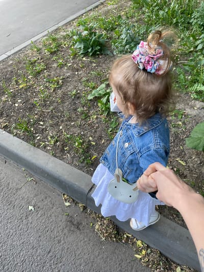 The image shows a young girl wearing a blue denim jacket, a white dress, and a white bunny purse, walking along a sidewalk in a city park. She is holding hands with an adult, who is out of frame. The girl is wearing a colorful patterned hair scrunchie, and she is wearing a medical face mask. The ground is covered in loose gravel and greenery. The scene is captured from a high angle, looking down. The coordinates provided (55.659, 37.591) place the scene in the vicinity of the city center of Moscow, Russia.