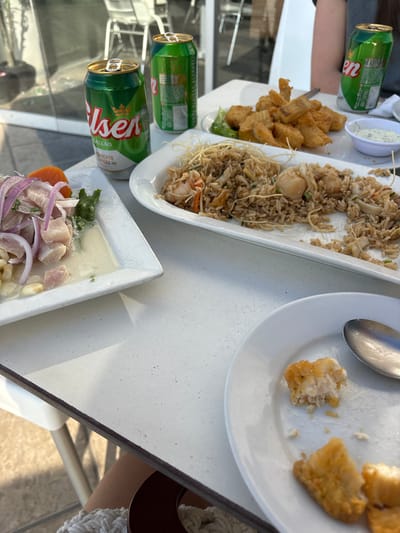 The image depicts a close-up, eye-level shot of a meal laid out on a white speckled outdoor table. Two green cans of Pilsen Callao beer are visible on the left, one opened. To the right of the beer cans, a rectangular white plate contains a portion of fried fish. Adjacent to this is a long white plate filled with fried rice, garnished with crispy noodles and containing pieces of seafood such as shrimp and scallops. In the foreground, a round white plate holds a small portion of fried fish, partially eaten, with a silver spoon resting beside it. Another white plate to the left holds a dish of ceviche, featuring chunks of white fish, red onion slivers, corn kernels, and a slice of orange. The setting appears to be an outdoor dining area, possibly a restaurant terrace, with hints of blurred outdoor seating and natural light suggesting a daytime meal. The overall scene conveys a casual dining experience.