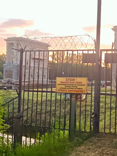 A rusty metal gate with barbed wire along the top is in the foreground. Behind the gate, a yellow sign with red and black lettering is attached to a metal pole. The sign reads: "СТОЙ! ЗАПРЕТНАЯ ЗОНА ТЕРРИТОРИЯ ОХРАНЯЕТСЯ СОБАКАМИ ПРОХОД, ПРОЕЗД, КУПАНИЕ, КАТАНИЕ НА ПОДКАХ СТРОГО ЗАПРЕЩЕНО!" This translates to: "STOP! PROHIBITED ZONE THE TERRITORY IS PROTECTED BY DOGS PASSAGE, TRAVEL, SWIMMING, RIDING ON TUBES IS STRICTLY PROHIBITED!"
Behind the gate and sign, a large, imposing concrete building with multiple windows is visible. The sky above is filled with soft, pastel-colored clouds, suggesting either sunrise or sunset. Trees and green grass surround the building and the gate. The overall scene appears to be in an industrial or fenced-off area within the city of Vodnik Snt, Russia.