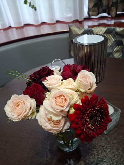 A vase of flowers, including blush roses, dark red roses, and a red gerbera daisy, sits on a dark wooden table. A decorative glass cylinder with vertical ridges is positioned behind the flowers. To the right of the flowers, a small clear glass cube holding a white candle is visible. The scene is set indoors, likely in a restaurant or cafe, given the presence of upholstered seating and curtains in the background, suggesting an urban setting. This setting could be Pila, Poland.