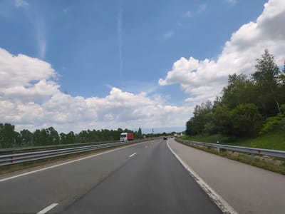 The image depicts a view from a vehicle on a highway, with the sky a vibrant blue dotted with fluffy white clouds. On the left side of the highway, a silver guardrail runs parallel to the road, with a dense line of green trees beyond it. A white truck with a red trailer is visible on the highway ahead. The right side of the highway is bordered by a grassy embankment covered in trees. In the distance, mountains are visible under the hazy sky. The road itself is paved asphalt with white lane markings. The overall scene suggests a sunny day on a rural highway. The location is confirmed as Malo Buchino, Bulgaria, by the context provided.