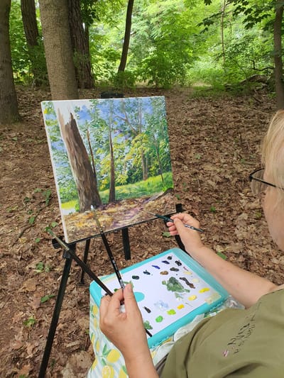 An outdoor scene in a wooded area depicts a person painting a forest landscape. The artist, viewed from behind and to the right, has fair-colored hair and wears glasses. They are holding a paintbrush and are focused on a canvas displayed on a tripod easel. The canvas shows a painting of trees with a central, prominent tree trunk, sunlight filtering through leaves, and a path. In front of the artist is a turquoise palette filled with various colors of paint, including greens, blues, yellows, browns, and white. Two paintbrushes are resting on the easel. The ground is covered with fallen leaves and some sparse vegetation. The background consists of dense green trees and foliage, suggesting a forest environment. The specific location in Falkensee, Germany is not discernible from the image.
