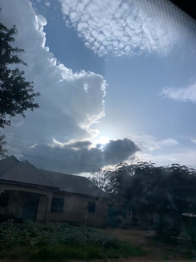 The image captures a view of Mangu Halle, Nigeria, featuring a partially obscured sun breaking through dramatic cloud formations in a bright blue sky. The sun's rays are visible, creating a light effect through the clouds. In the foreground, a low-lying building with a corrugated metal roof is visible, appearing weathered and aged. Adjacent to the building is dense green foliage. Several power lines are strung across the scene, running horizontally in the middle ground. The overall impression is of a rural or semi-rural environment under a dynamic sky.
