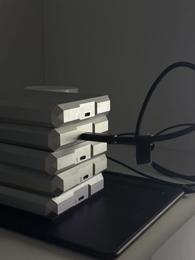 An indoor, low-light photograph captures a collection of electronic devices arranged vertically. Five silver-colored, hexagonal prism-shaped objects, possibly external hard drives or small form-factor computers, are stacked or lined up. A black power or data cable descends from above, secured by a black hook-and-loop strap, and connects to one of the units. Other black cables are visible in the dimly lit background. To the right, a dark, rectangular electronic device, likely a tablet, leans against a lighter surface. Each hexagonal unit has a small rectangular port on its top surface, accompanied by a numeric label. From left to right, the clearly visible labels on the units are '4', '3' (the unit into which the cable is plugged), '2', and '5'. The overall scene suggests an organized setup of electronic equipment in a workspace or server area, under low ambient lighting. The location is Башкултаево, Russia.