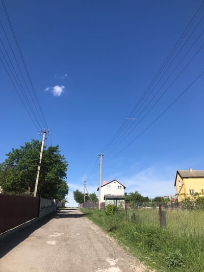The image shows a dirt road in Petrykiv, Ukraine. The road is bordered by fences, vegetation, and houses. Overhead power lines extend across the clear blue sky. No people are visible.