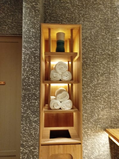 The image shows a wooden shelving unit with three visible shelves, located in Kendhoo, Maldives. The shelves are illuminated from within and display rolled white towels. The top shelf holds a single decorative object, a cylindrical vase with a green band. The shelving unit is positioned against a wall with a speckled, textured finish. To the left of the shelving unit, a portion of a door is visible. The environment appears to be a modern, minimalist interior, possibly a spa or bathroom.