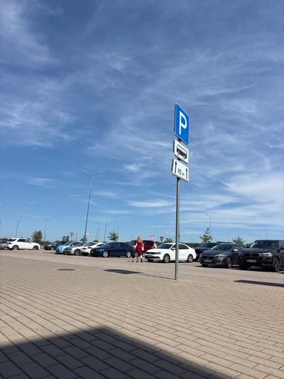 A bright, outdoor, daytime shot captures a parking lot with a clear blue sky and scattered white clouds. A tall metal pole with a parking sign, a bus symbol, and a sign indicating "90 m" with arrows stands prominently in the foreground, slightly right of center. The parking lot pavement is made of light grey interlocking bricks. Several cars are parked in rows. On the left side, a white SUV and several sedans of various colors, including blue, silver, and dark grey, are visible. On the right side, a dark grey SUV is parked.
Two individuals are walking in the parking lot. A woman in a red dress is walking towards the right, and a shorter person, possibly a child, is walking beside her. In the background, a row of trees and more streetlights can be seen. The scene appears to be in a parking lot located in the city of Piņķi, Latvia.