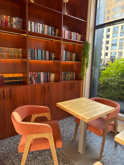 This is a photograph of a modern, well-lit indoor space that appears to be a library or study area within a building. The primary subjects are a large wooden bookshelf filled with books, two modern orange chairs, and a light wood table.
The bookshelf, made of dark wood, is organized into multiple rectangular shelves, each densely packed with books of various sizes and colors. Some shelves are illuminated by subtle LED strip lighting, creating an inviting atmosphere. A small green plant is placed on top of one section of the bookshelf.
The chairs are contemporary in design, with curved orange upholstered seats and backs, supported by light wood frames. One chair is placed at the table, while another is positioned to the side, its back facing the camera. The table has a light wood, angular top and a grey metal pedestal base.
In the background, a large window offers a view of an urban environment. A multi-story building with numerous windows is visible, along with lush green foliage, suggesting it is daytime and likely a pleasant day outdoors. The floor is a speckled terrazzo material.
The scene conveys a sense of quiet study or relaxation. The lighting is bright and even, indicating the photograph was taken during daylight hours. The visible text on the book spines includes titles and author names in Cyrillic script, suggesting the location is likely Russia. Specific visible text includes "ВАНКИР", "СТАЛИНГРАД", "ДАНТЕ АЛИГЬЕРИ", "ДУГЛАС АДАМС", "1984", and "ГЕРМАН ГЕСС".