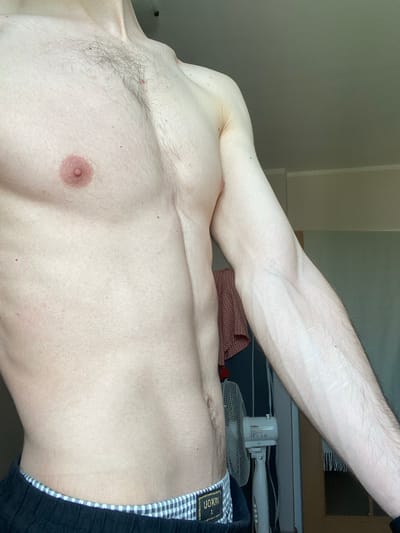 The media file shows a close-up, horizontal shot of a person's torso and arm. The person is fair-skinned and appears to be male, with a visible nipple on the chest and some chest hair. The arm is extended and raised, revealing prominent veins. The person is wearing black and white checkered boxer briefs with a visible brand tag. In the background, a small, white electric fan is visible, along with a patterned fabric and what appears to be a desk or surface with striped material. The lighting suggests an indoor setting, possibly a bedroom, and there are no clear indicators of the time of day or weather. The overall impression is a casual, personal snapshot. There is some text visible on the brand tag of the boxer briefs, which appears to read "UOKIN".