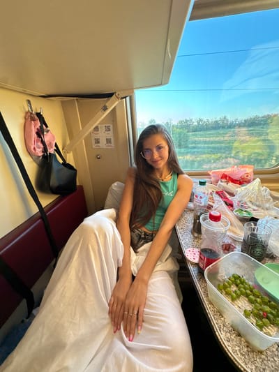 A young woman with long brown hair and glasses is seated in a train compartment. She is wearing a turquoise tank top and dark pants. A white blanket is draped over her legs. On the table in front of her are various food and drink items, including a container of green berries, bottles of water and soda, and bags of snacks. The train compartment appears to be in motion, with scenery visible through the window. The overall impression is of a casual journey. There is no indication of the city of Фисенково, Russia in the image.