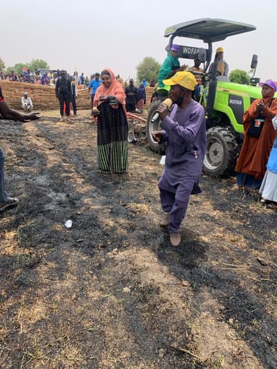 The image shows a scene in Ringimawa, Nigeria (latitude: 11.666, longitude: 8.416). The ground is covered in black ash and there are numerous people standing around. One man wearing a blue shirt and a yellow hat is speaking into a microphone while facing the camera. He is standing in front of a green tractor that is parked on a path. To his left, a woman is holding her hands together. There is a large group of people in the background, including some men wearing red shirts. There are also some stacks of sticks in the background. There are trees in the background and a clear sky above. The scene suggests a gathering of people, potentially for a community event or a political rally.