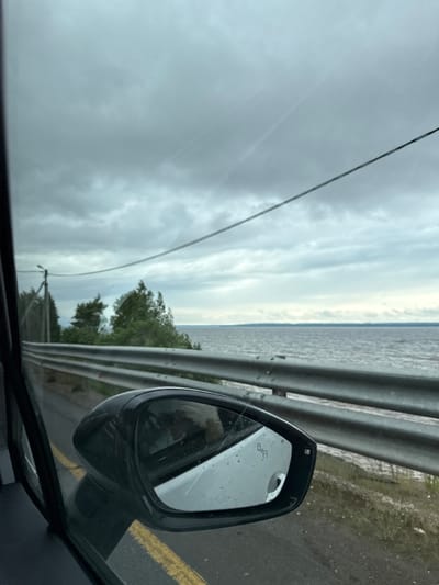 The media file displays a roadside view from within a vehicle, capturing a landscape in what appears to be Kryukovo, Russia. The scene depicts a large body of water, likely a lake or sea, stretching towards a distant horizon under a heavily overcast, grey sky.

In the foreground, the black side mirror of the car is prominently visible on the right, showing reflections and a symbol for blind-spot monitoring (a small car with a diagonal line through it). The road's edge features a yellow painted line, and a multi-bar metal guardrail runs horizontally along the roadside, separating it from the water. Between the guardrail and the water, a scattering of green trees and foliage is visible. A thin power line or utility wire stretches across the upper left portion of the cloudy sky.

The overall impression is that of a daytime scene on an overcast day, possibly post-rain or during damp weather, given the appearance of the road surface near the guardrail. The water surface shows ripples, indicating movement or wind. The activity implied is traveling or being parked by the water.
