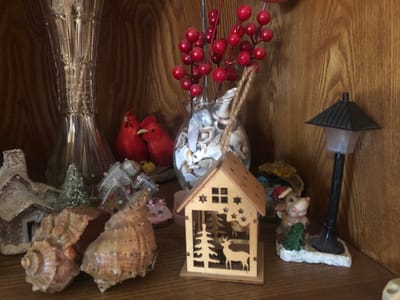 The image displays a collection of decorative items on a wooden shelf, likely in a home environment. The arrangement includes several miniature houses, some appearing to be decorated as gingerbread houses, and a small wooden house with a Christmas tree and deer cutouts. There are also seashell decorations, a glass vase filled with shells and red berry branches, and two red ceramic roosters. A small figurine of a cow wearing a Santa hat sits near a miniature lamppost. The background is a wooden paneled wall. There are no people present, and no actions or interactions are depicted. The scene is static and focuses on the decorative objects. The image does not provide any information about the city of Chiatura, Georgia.