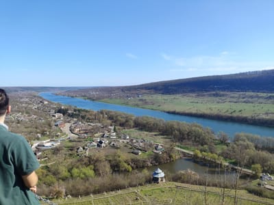 A daytime, eye-level, medium shot captures a picturesque view from Stroiesti, Moldova. A river meanders through a valley, bordered by a village and a wooded area. The sky is a clear, vibrant blue. A man in a green shirt stands at the left edge of the frame, observing the landscape. A small pavilion structure is visible near a body of water in the foreground, along with trellised plants.