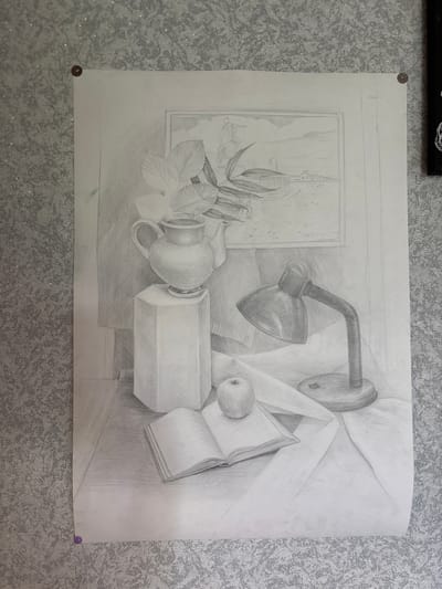 This is a pencil sketch of a still life, featuring a lamp, a pitcher, an apple, an open book, and a rectangular pedestal, arranged on a surface draped with fabric. A framed picture hangs on the wall behind the arrangement, depicting a landscape with a pier and buildings. The sketch is tacked to a textured, sparkly wall, which is a common feature in homes. The location of Ушаки, Russia is irrelevant as the image only depicts an interior scene and provides no geographic information.