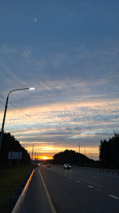 This media file shows a highway scene at sunset, likely in Bogandinsky, Russia. The viewpoint is from the side of the road, looking forward along the lanes. The sky is filled with dramatic clouds, illuminated with warm hues of orange, yellow, and pink from the setting sun, contrasted with areas of deep blue. Wispy, streaky clouds are visible throughout the sky, with some appearing more textured and broken.
On the right side of the image, the asphalt road is marked with white dashed lines and a solid yellow line. A silver guardrail runs along the edge of the road. Several cars are visible driving on the highway, their headlights on, indicating it's dusk. One car in the foreground is a dark sedan, and another further down the road appears to be a similar model.
The left side of the highway is bordered by a dense line of dark green trees, suggesting a forested or wooded area. Tall streetlights with white glowing lamps stand along the roadside, one prominent in the foreground. Power lines stretch across the sky. A road sign is partially visible in the lower left, but any text on it is illegible.
The overall mood of the image is peaceful and serene, capturing the beauty of a sunset over a transportation artery. The time of day is clearly sunset, and the weather appears clear with dynamic cloud formations. The scene conveys a sense of travel and the transition from day to night.