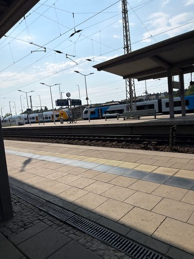 This media file captures a scene at a train station in Eberswalde, Germany. The perspective is from a train platform, looking towards the tracks and oncoming trains. The platform itself is paved with light-colored square tiles, with a textured yellow stripe running parallel to the tracks, likely a safety marking. To the right of the platform, a drainage grate is visible.
Several trains are present, including a blue and white train parked at the platform, and a yellow and white train further down the tracks. The trains appear to be modern passenger trains. Overhead, a complex network of electrical catenary lines and support poles stretches across the sky, indicating an electrified railway. The sky is blue with scattered white clouds, suggesting it is daytime, likely late morning or afternoon given the strong shadows cast on the platform.
Notable details include a bicycle parked on the platform next to the yellow and white train, and a clock face visible on a pole in the distance. There is also a digital display board providing information, though the text is too small to be legible. One of the white trains has "odeg.de" visible as text, which is the website for the Ostdeutsche Eisenbahn, a regional railway company.
The scene depicts a typical operational day at a train station, with no specific activity or event highlighted beyond the presence of the trains and the infrastructure. The overall impression is one of orderly transit and daily life.