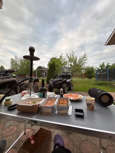 The image depicts a picnic or outdoor meal set on a dark grey folding table, likely in a yard or garden. The location appears to be Ogryzkovo, Russia, based on information not visible in the image. The table is covered with food items in various containers, including a plate of slices, several foil containers with cooked food, and food in open cardboard boxes. A speaker, a patterned paper cup, and a small black container are also on the table. A hookah stands on the table, with its mouthpiece facing the viewer.

In the background, there are two all-terrain vehicles (ATVs) parked on the grass. Beyond the ATVs, there are trees, a trampoline, and what appears to be a building or house. The sky is cloudy. Parts of footwear, possibly a slipper, and what looks like a brown bag are visible on the right side of the table. The table legs are visible on the right. The scene indicates a casual gathering or meal outdoors.