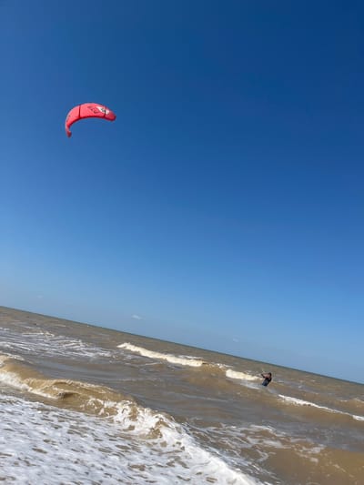 A red kite is visible in a clear blue sky over a choppy, brown sea. Below the kite, a person is kitesurfing in the waves. The scene appears to be at a beach in Dolzhanskaya, Russia, with the water meeting the shore at an angle.