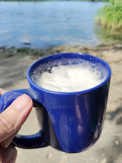 A person's hand holds a blue mug filled with a frothy white beverage, likely coffee or a similar drink, outdoors. The mug is positioned in the foreground, obscuring the lower portion of the scene. The background shows a body of water, possibly a lake or river, with a shoreline featuring sand and sparse vegetation. In the distance, the water reflects the blue sky, suggesting a bright, clear day. The sunlight creates dappled shadows on the ground. The overall impression is one of a relaxed moment, enjoying a drink in a natural setting. The image was taken in San Martín de Porres, Peru. There is no visible text in the image.