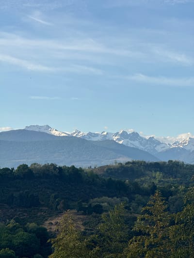 The image shows a landscape view, likely taken in a mountainous region. The sky is predominantly blue with some wispy clouds on the left side of the image. In the center, a mountain range is visible, some of its peaks capped with snow. To the right of the mountains, there is a dense green forest covering the lower slopes. The photograph appears to be taken during daylight hours. The exact location within Tsanbaikwara, Abkhazia is not possible to determine from the image alone. There are no people visible in the photograph.