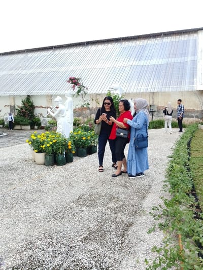 The image depicts an outdoor scene in what is stated to be Ngadirejo, Indonesia. A group of three adult women occupies the left-center foreground on a light-colored gravel path. The woman on the left, wearing a black long-sleeved shirt and sunglasses, holds a smartphone vertically. The central woman, in a red top and dark capri pants, has her left arm around the shoulder of the woman to her right, who wears a light blue denim-style dress and hijab. Her right hand is extended towards the phone. To the left of these women stand two white classical-style female statues, one appearing to play a wind instrument. Adjacent to the statues are several potted plants with vibrant yellow flowers, some in white pots, others in dark green grow bags. The foreground path is bordered on the right by a narrow strip of grass lined with small plants in black containers. In the mid-ground, a woman in a hijab and dark top, and a man in a plaid shirt, are visible standing on a separate, unpaved area. The background features a large, weathered building with an off-white lower wall and a corrugated metal or translucent roof. The sky is overcast.