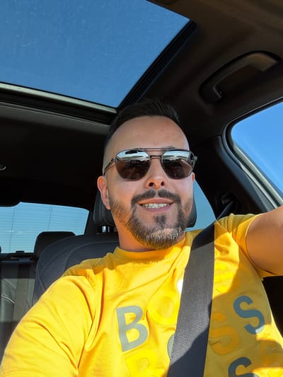An adult male with dark hair, a beard, and aviator sunglasses is smiling while taking a selfie from the driver's seat of a vehicle in Kolarovo, Bulgaria. He is wearing a bright yellow t-shirt with repeated 'BOSS' lettering, some in gray and some in yellow, and a black seatbelt is visible across his chest. The car features a dark interior and a panoramic sunroof, through which a clear blue sky is visible. Reflections in his sunglasses show an open, sunlit landscape. The scene is brightly illuminated by natural light, indicating daytime.