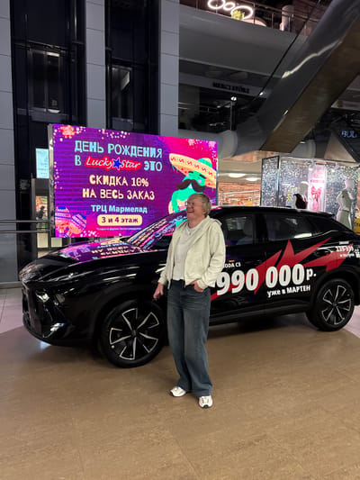 A woman stands in front of a black car with promotional graphics in a shopping mall. The woman is wearing glasses, a light-colored hooded jacket, and blue jeans. She is smiling and looking to her right, with her left hand in her jeans pocket. The car has large red graphics on its side, including the text "990 000 p." and "уже в МАРТЕН." A large digital screen behind the car displays promotional text in Russian, including "ДЕНЬ РОЖДЕНИЯ В Lucky Star ЭТО СКИДКА 10% НА ВЕСЬ ЗАКАЗ" and "ТРЦ Мармелад 3 и 4 этаж". The mall environment is visible in the background, with escalators and storefronts. The floor is tiled.